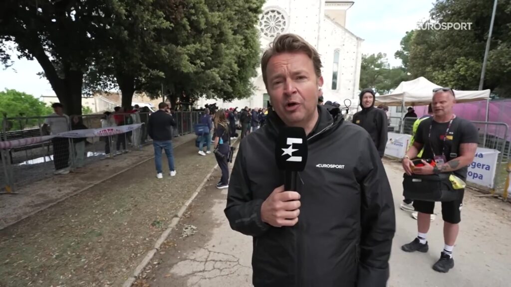 Een Eurosport-verslaggever met microfoon bij de start van een Giro-etappe in 2026, uit een aflevering van Kop over Kop